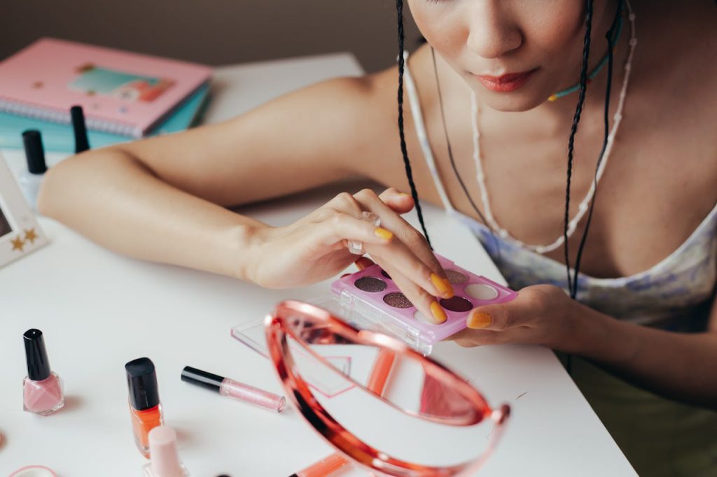 pexels photo 10608368 Woman using eyeshadow palette with cosmetics scattered on a table, reflecting in mirror.